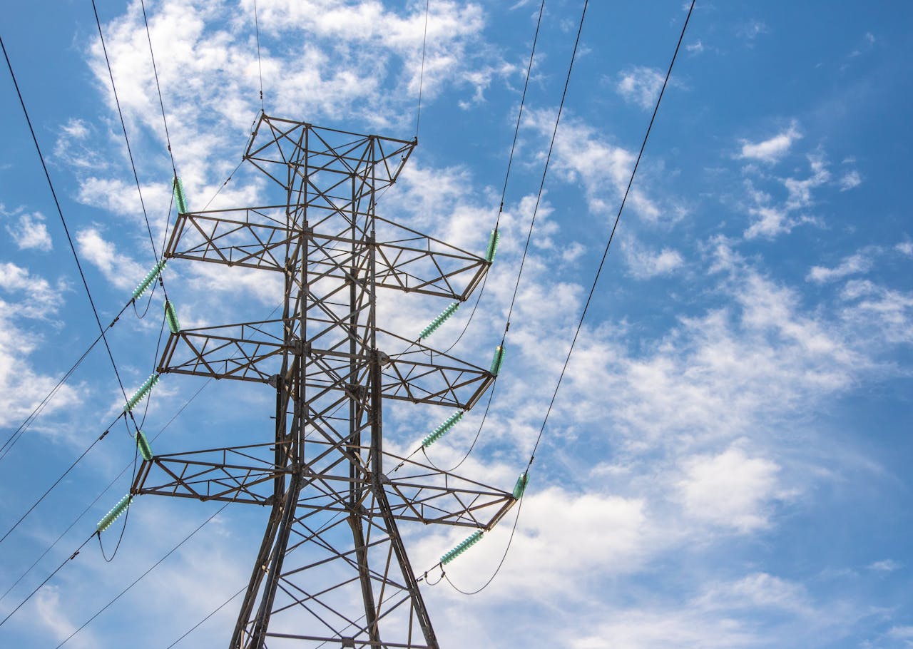 High voltage power line tower framed against a bright blue sky with clouds.